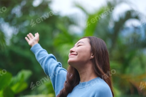 Preview: Portrait image of a woman with closed eyes raising hand while standing in the garden