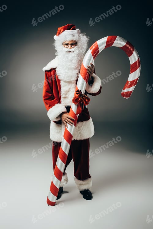 Preview: happy santa claus in costume standing with big striped christmas stick on grey background
