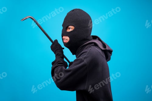 Preview: Portrait of Sneaky Masked Criminal Using Crowbar Isolated Over Blue Studio Wall