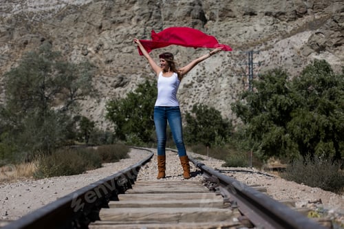 Visualização: jovem mulher bonita em pé em uma ferrovia e brincando com um xale vermelho. Ao ar livre. Ensolarado. Lifestyle