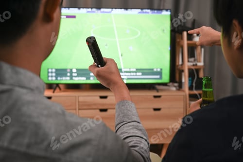 Preview: Two casual men cheering football match while sitting on sofa in living room.