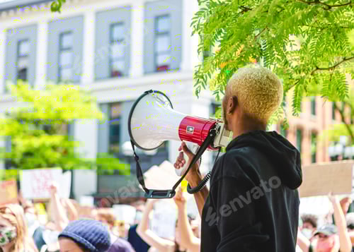 Preview: Person of color holding a megaphone at a peaceful protest, speaking to a crowd of activists w/ signs