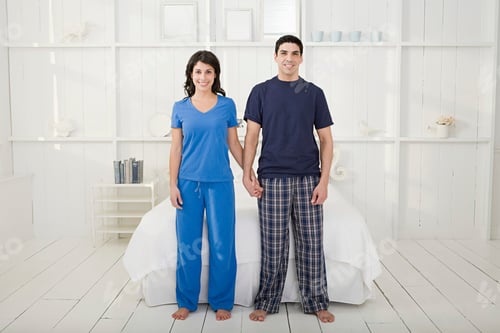 Preview: Couple in sleepwear holding hands with a bright smile in a cozy, white bedroom setting.