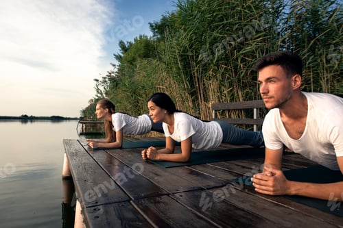 Preview: Group of people doing yoga exercises by the lake at sunset.