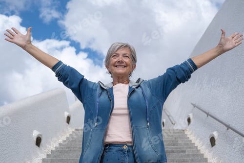 Preview: Happy adult mature senior woman with open arms outdoors on staircases looking away smiling