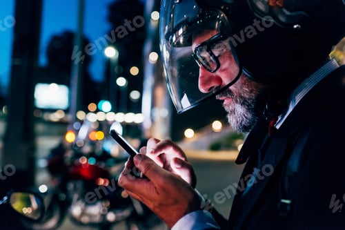 Preview: Mature businessman outdoors at night, wearing motorcycle helmet, using smartphone, profile