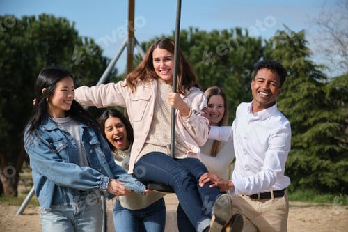 Preview: Group of happy young adults having fun pushing a woman on a swing
