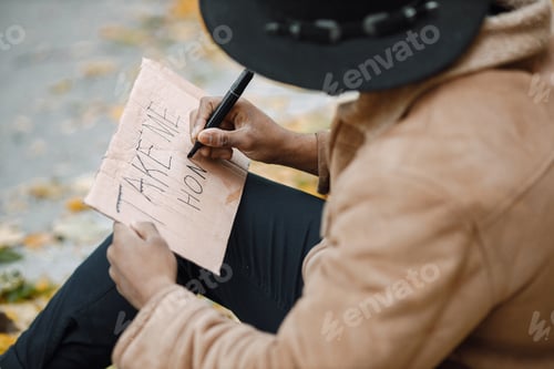 Preview: Black man sitting on a road and writing a sign for autostop