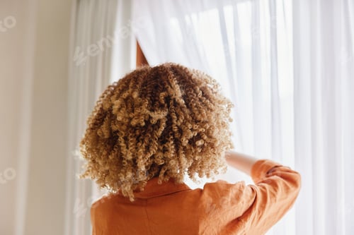 Preview: A young woman with curly hair adjusts sheer curtains, creating a warm and inviting atmosphere in a