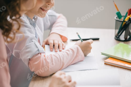 Preview: cropped view of mother and daughter sitting at table and doing schoolwork together at home