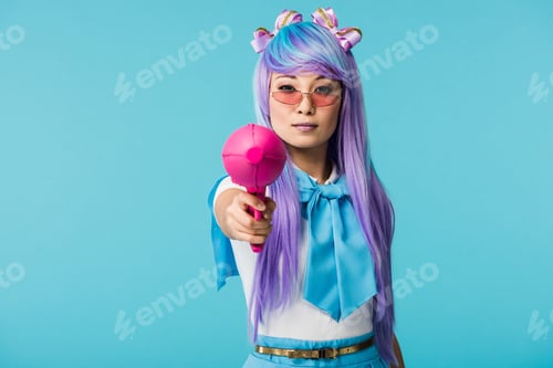 Preview: Serious asian anime girl in wig and glasses holding water gun isolated on blue