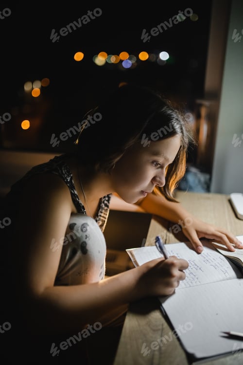 Preview: high school girl doing homework at the table in a dark room, close-up.
