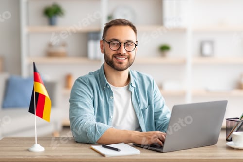 Preview: Happy young guy sitting at table with flag of Germany, using laptop pc, studying foreign language