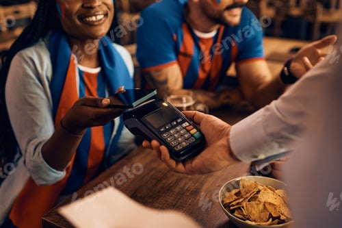 Preview: Close up of black woman paying contactless during sports championship in a bar.