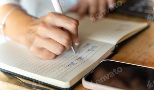 Preview: Close-up view of a woman writing on a notebook while working with a laptop in a coffee shop.