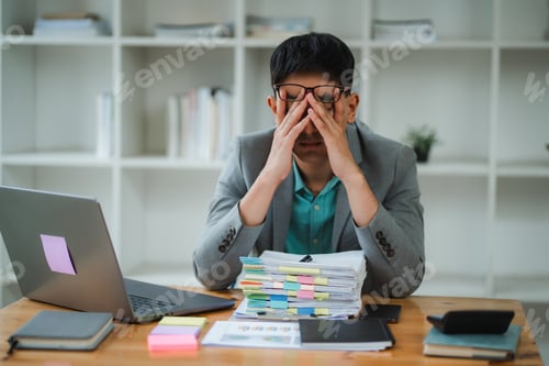 Preview: Stressed asian businessman covering face with hands at office desk