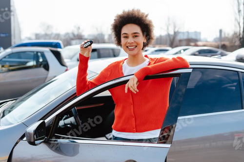 Preview: Beautiful, smiling African American woman, happy female owner holding car key standing near car