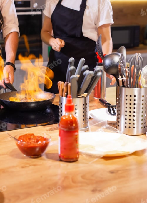Preview: Female Chef in restaurant kitchen at stove with pan doing flambe on food