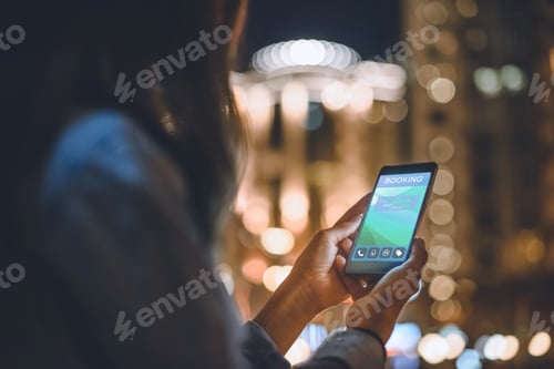 Preview: partial view of woman using smartphone with booking logo on screen night city lights on background