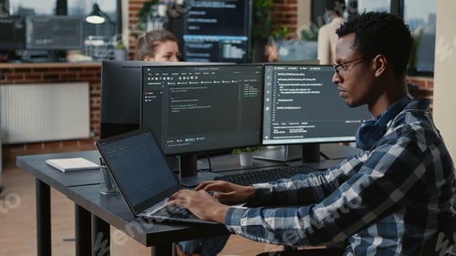 Preview: Portrait of tired programer rubbing eyes while typing on laptop sitting desk with multiple screens