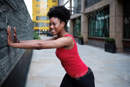 Preview: Portrait of smiling young fit black woman stretching against a wall.
