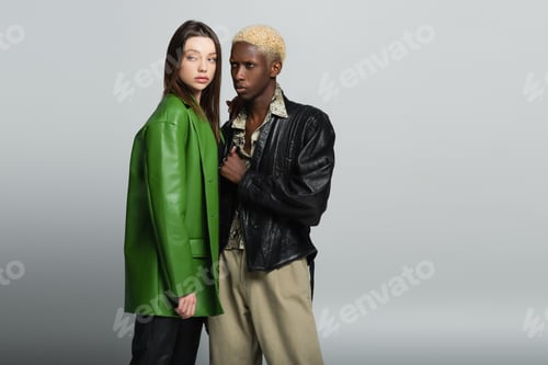 Preview: blonde african american man and brunette woman in leather jackets standing on grey