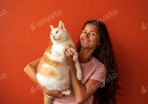 Preview: Happy young brunette caucasian woman holding her white and red cat on orange background.
