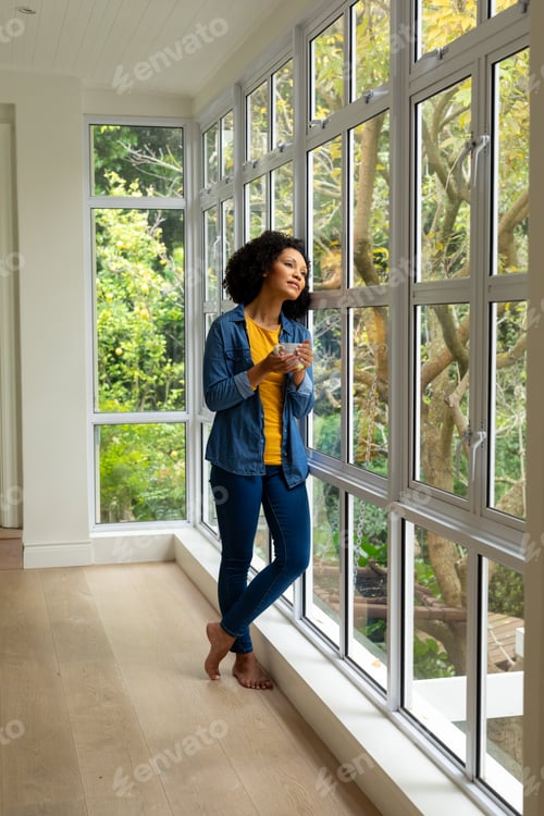 Preview: Biracial woman holding cup of coffee and looking out window at home