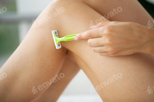 Preview: Young woman removing hair on legs with razor at bathroom.
