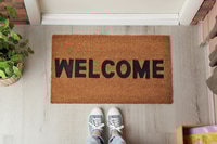 Preview: Woman standing near door mat with word Welcome on wooden floor in hall, top view