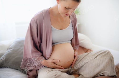 Preview: Happy pregnant woman stroking her belly, sitting on bed.