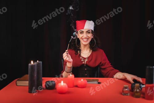 Preview: Cheerful gypsy soothsayer holding match near candles and blurred witchcraft supplies isolated on