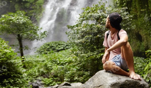 Preview: Outdoor shot of stylish young Caucasian hiker sitting barefooted on big rock and looking over his sh