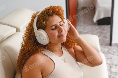 Preview: Curly-haired woman enjoying music with headphones, smiling peacefully on a sofa at home
