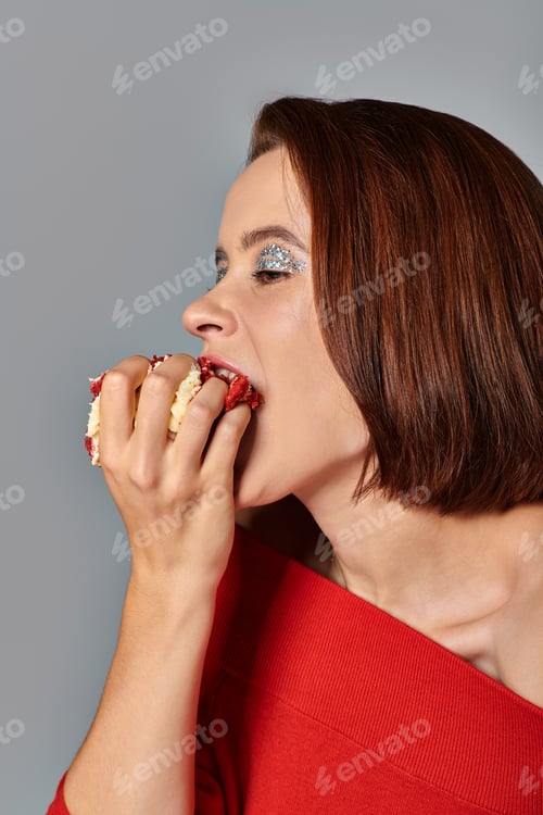 Preview: beautiful young woman in red attire biting delicious piece of birthday cake on grey background
