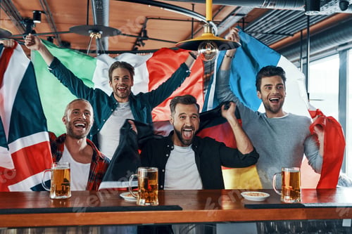 Preview: Excited young men covered in international flags drinking beer and watching sport game