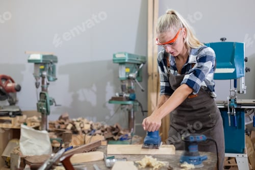 Preview: young carpenter caucasian woman using electric wood sander on wood at manufacturing wooden industry