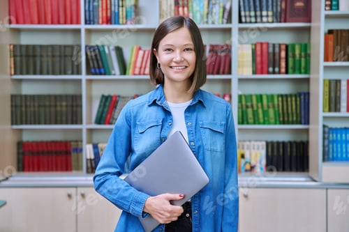 Preview: Young female university student looking at camera, standing with laptop in library