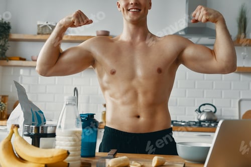 Preview: Unrecognizable fit man showing his biceps and smiling while preparing healthy food at the kitchen