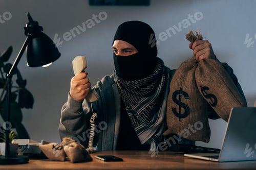 Preview: Aggressive terrorist in mask holding money bags and looking at handset