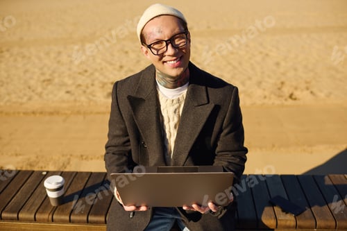 Preview: Stylish young man with tattoos working on laptop at sandy beach under bright sun
