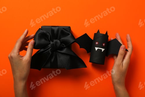 Preview: Gift, paper bat and hands on orange background, top view