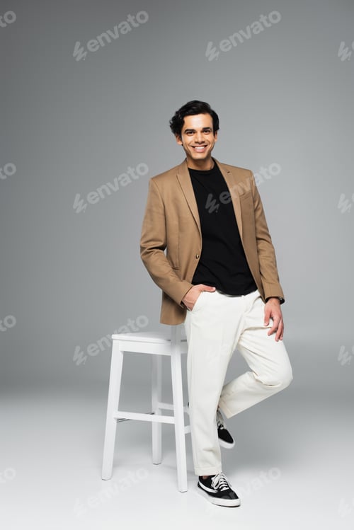 Preview: full length of happy young man in blazer standing with hand in pocket near white chair on grey