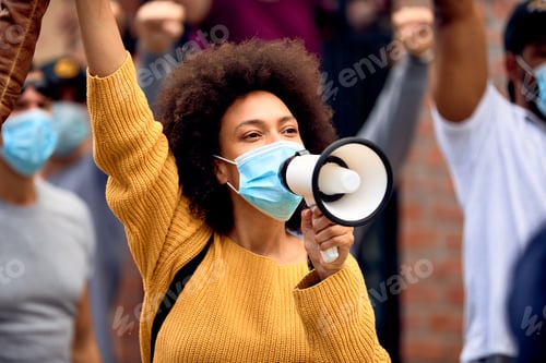 Preview: Black female activist wearing protective face mask while shouting through megaphone on a protest.