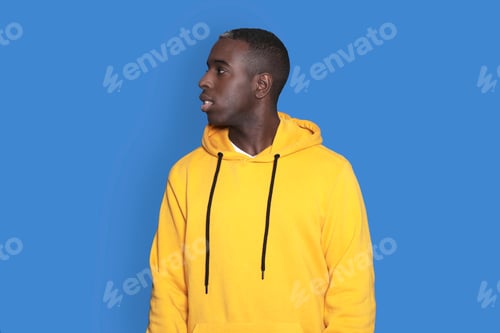 Preview: young african-american guy wearing yellow hoodie posing isolated on blue background looking aside