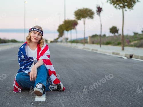 Preview: Blonde Caucasian woman from Spain sitting on a road smiling with a USA flag over her shoulders