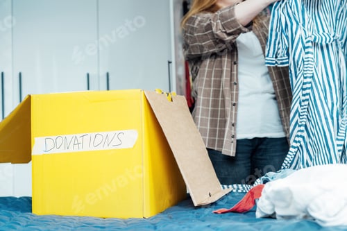 Preview: Yellow cardboard box of selected clothes for donating to a Charity shop standing on the bed.