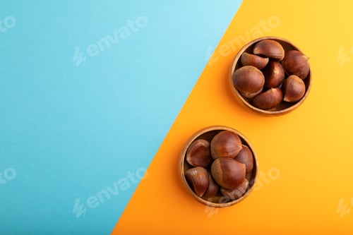 Preview: wooden bowl with raw edible Сhestnuts on blue and orange paper pastel, flat lay, copy space