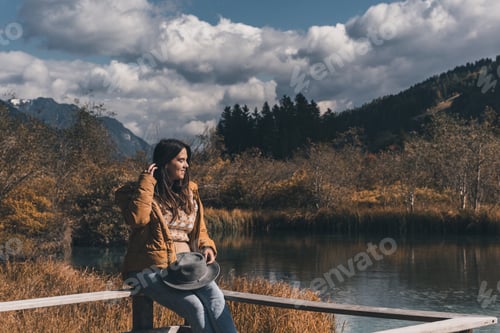 Preview: Beautiful young woman wearing autumn clothes, enjoying view of moody fall scenery by a lake