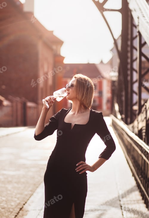 Preview: Attractive happy girl in a black dress walks along a city street with a glass of wine.